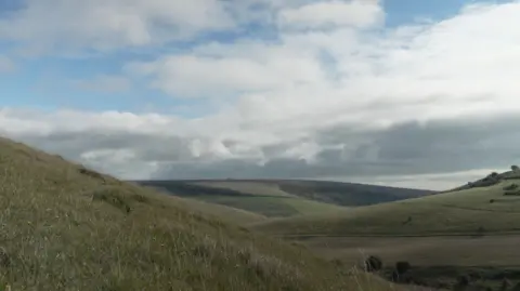 Kevin Church/BBC News A picture of the South Downs with rolling hills off into the distance and a mostly cloudy sky above with a few patches of blue. 