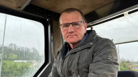 A farmer, a man with short black hair wearing glasses and a coat, sitting in the cab of a tractor on a rainy overcast day.