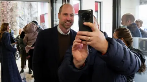 PA Media The prince smiling while looking into a mobile phone which a person with a blue jacket on is holding up next to him.