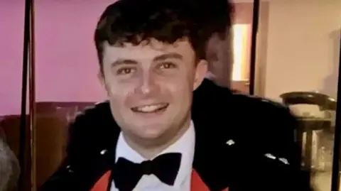 MOD Captain Philip Gilbert Muldowney smiles as he looks into the camera, with short dark hair, wearing a dress suit and bow tie.
