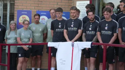 Players and staff of Scunthorpe United wearing training tops and shorts gather outside the club's ground with their heads bowed. On the railings in front is a white England shirt with the words "RIP legend Kevin Keegan".