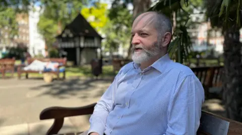 Conservative candidate Tim Barnes, who has a grey beard and is wearing a shirt and sitting on a bench