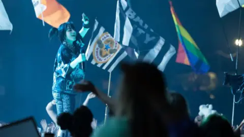 Getty Images Billie Eilish stands on the Pyramid Stage at Glastonbury, singing into a microphone and holding her hand up as flags being held up by the crowd surround her. She is wearing a baggy black T-shirt with white patterns on it, and shorts of a similar style. He black hair is in bunches. The silhouettes of the crowd can be seen in the foreground.