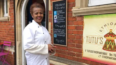 BBC A lady in chef's whites stands outside her cafe, in a red brick Victorian building.  She is smiling.  There is a pink metal table and chairs behind her.  Over her shoulder is a blackboard advertising their social media contacts.  To the right of the picture is the sign for the cafe - you can just make out the word 'Tutu's'.