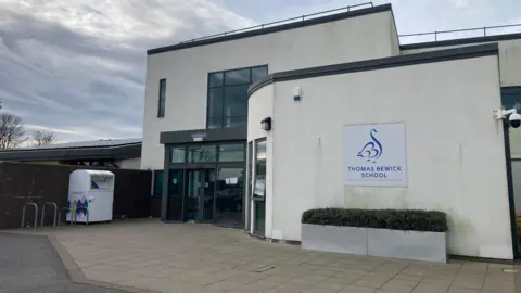 The entrance to Thomas Bewick School which is a small building with white concrete walls and windows painted brown. To one side are bike racks and a recyling container