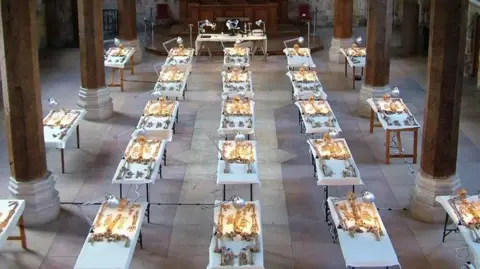 York Archaeology Twenty one tables set out in a large hall all which have sekeletal remains on display. The tables are set apart from each other and are all lit up by a lamp.