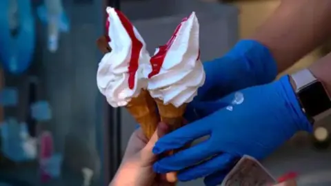 Getty Images A close up of a pair of hands in blue catering gloves handing over two whippy ice cream cones with raspberry sauce