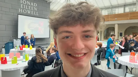 A teenage boy wearing a grey shirt and black blazer. He has short, curly brown hair. 