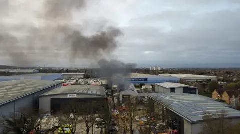An aerial shot of the industrial estate, showing smoke coming from the roof of one building. There are many large warehouses around and emergency vehicles next to it.