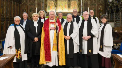 Keith Mindham The group of male and female senior clerics dressed in white and black robes, with clerical collars. Archdeacon Brazier-Gibbs stands to the side of Bishop Graeme Knowles, who is holding a crosier, and wearing a white robe underneath a gold and yellow cape. They are standing in front of an altar, surrounded by wooden carved fittings and fixtures and religious paintings.