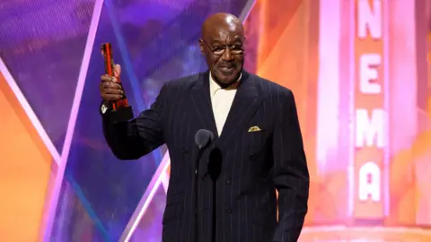 Delroy Lindo accepting an AARP award wearing a pin-striped dark jacket and white shirt
