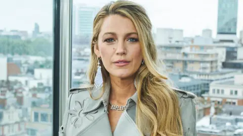 Blake Lively is wearing a grey jacket and stood with a London skyline behind her.