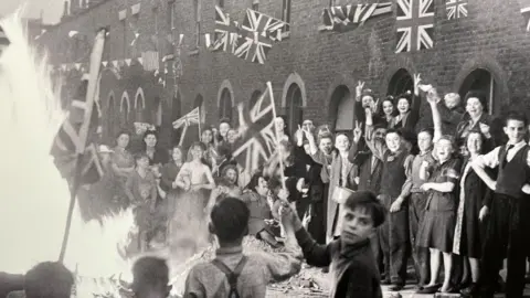 People laughing and smiling while celebrating VE Day in a London Street that has been bombed. There is a bonfire in the street and there is Union Jack bunting above their heads and a child with his face turned to the camera is waving a flag on a pole.