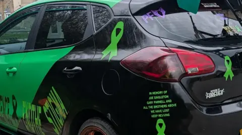 Ben Drew The rear end of the car which is painted black. It has a lot of green mental health ribbons with a tribute to his two friends and "all the brothers above we have lost" written in green. 