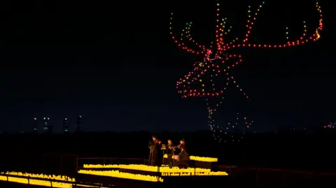 Fever A string quartet are surrounded by yellow candles while, overhead, drones lit up in the night sky make out the shape of a deer with antlers.