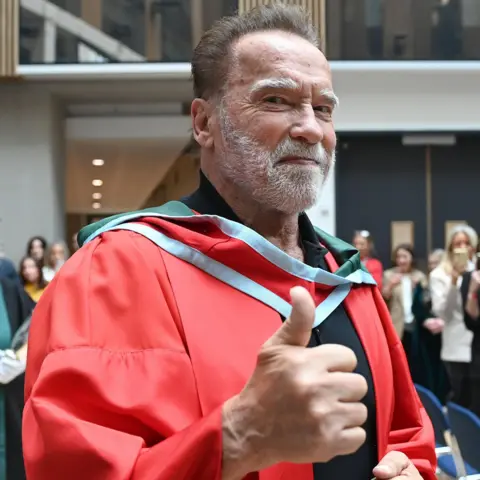 Getty Images Arnold Schwarzenegger has dark hair and a white beard. He is smiling and giving thumb's up. He is wearing a red university gown with a white and red collar