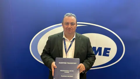 Isaac Chenery/BBC A man with short dark hair wearing a green blazer and a white shirt standing in front of a blue backdrop holding a folder which reads Maritime Transport