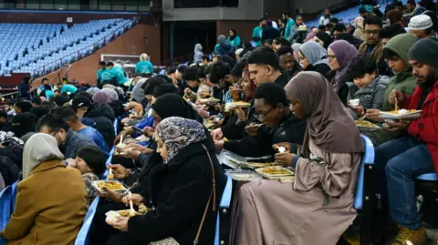 Dozens of people gather to break their fast together with a meal in the stand of a football stadium.