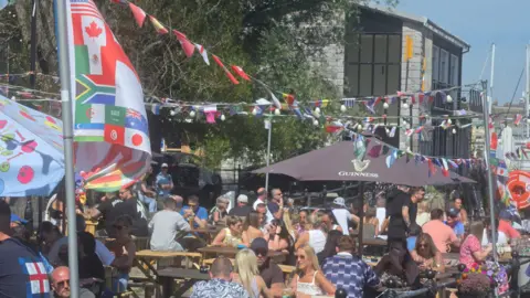 A picture of a number of people sat in a pub garden. There is a number of flags pictured above the tables.