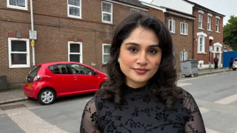 Martin Heath/BBC Alia Khan with shoulder-length dark hair stands on a residential street in front of a row of brick terraced houses. The person is wearing a black semi-sheer top with a floral pattern and pearl-style earrings. A red hatchback car is parked along the roadside behind them.