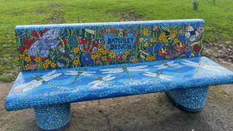 A blue bench in a park made up of tile pieces. In the centre it says Batchley bench