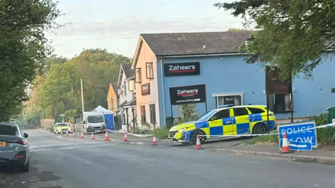 Two police cars and police tape around a row of three buildings next to a road. 