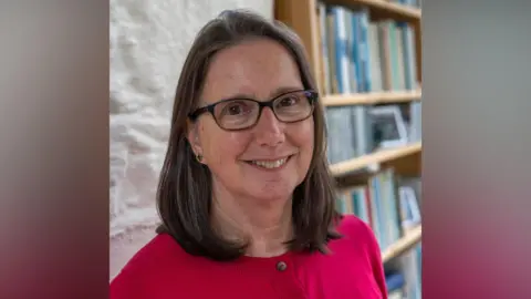A woman with a pink top, dark hair and glasses smiling at the camera.