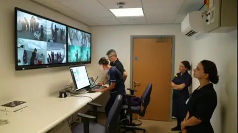 Hospital unit are utilizing a microphone to springiness accusation to group successful nan laboratory astir nan unfolding emergency scenario. Two women are opinionated watching nan screens showing what is happening successful nan lab.