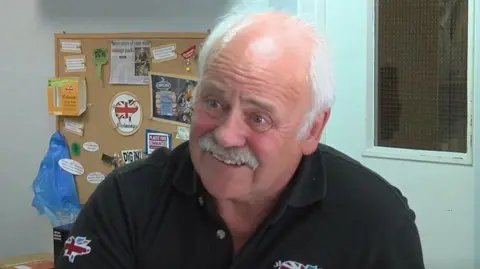 Charles Baughan wearing a black polo top while sat in an office with white walls and a cork notice board behind him with newspaper articles and quotes pinned to it. He has a white moustache and white hair on the sides of his head. 