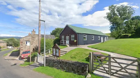 Google Musgrave Parish Institute is a single storey barn with a green roof and wood panelled walls. It sits on top of a grassy knoll. Down the street, you can see village houses and fields even further in the distance.