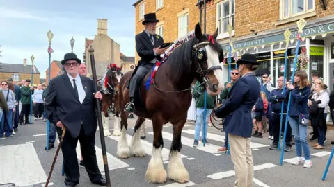 Carroll Weston/BBC The town bailiff on horseback in the street, reading from a book. People holding long blue poles with yellow diamond shaped tips surround him.