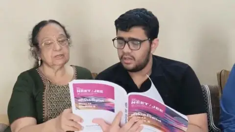 ANI An elderly woman and a young man wearing glasses sit together indoors, holding NEET/JEE preparation books and reading from them.