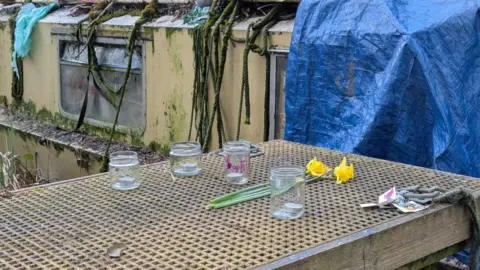 Arranged on the wooden jetty to the narrowboat are four tea lights in jam jars, a small bunch of daffodils and some drawings.