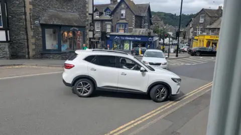 Ambleside The Wrong Way A white car turning the wrong way onto a one-way street in Ambleside, Cumbria, obstructing another white car which is travelling the correct way. There are shops in the background.