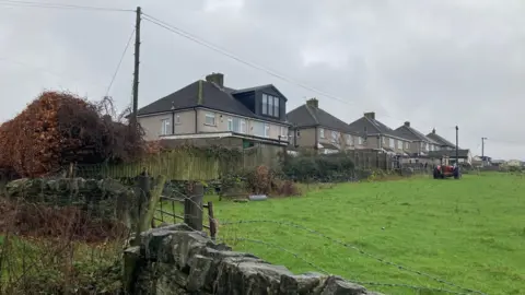 A row of semi detached houses backing onto a green field with an old red tractor in it 