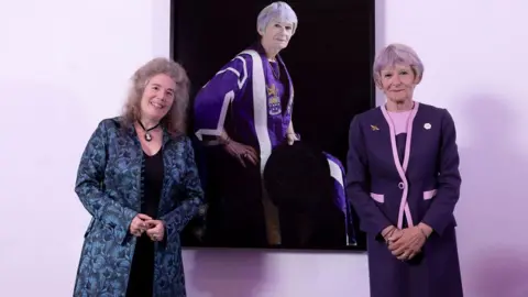 David Oates/UoM Photographer Dr Carla van de Puttelaar, smiling with greying hair and wearing a blue and black floral jacket, and Prof Dame Nancy Rothwell, smiling with grey hair and wearing a pink-trimmed purple jacket and skirt, stand in front of a portrait of Dame Nancy wearing her purple and silver ceremonial robes