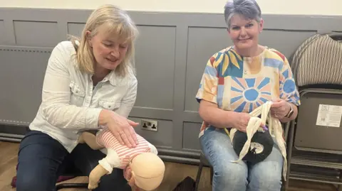 BBC/Katie Radley Two women are sitting on chairs in front of grey panelling. One woman is holding a mannequin upside down on her forearm with the other hand on its back. The other woman is tying a bandage around a cushion on her knee.