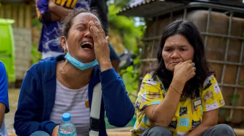 A woman wearing a white and pink shirt with a blue cardigan weeps, placing one hand over her eye, while another woman watches on 