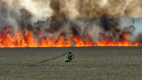 A firefighter dragging a hose across a dry field. In the background is a wall of flames with thick black smoke rising