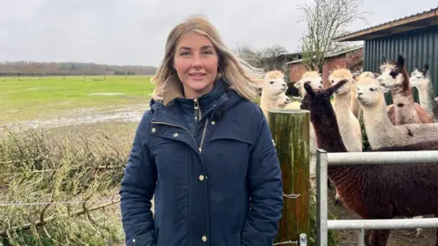 A photo of Natalie Neadley smiling into the camera. She is wearing a navy coat and has long blonde hair. Christmas trees lay on the grass behind her on the left and on the right is a herd of alpacas.