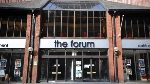 A general view of entrance to The Forum in Barrow, Cumbria. The signs above the glass doors and windows are black and white. There are show posters on the windows.