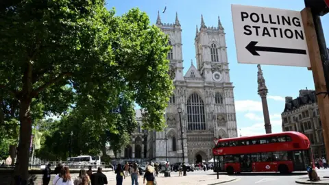 Westminster Abbey on a sunny day. There is a red London bus driving past the abbey and a sign affixed to a post that says Polling Station. 