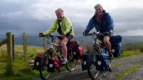 Fiona, on the right-hand side, is cycling on a bike covered in panniers next to her partner, Andy Blance, on the same set up, on a gravel path.