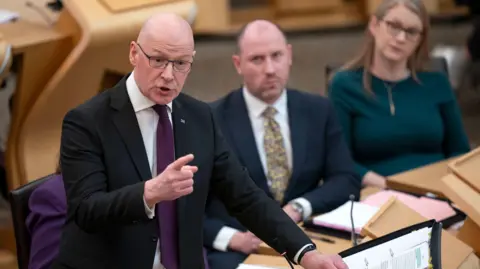 Pa Media John Swinney berdiri di sebuah meja di Kamar Holyrood