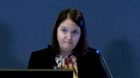 Omagh Bombing Inquiry Ms Travis has short brown hair and is wearing a black blazer. She has an expressionless face.