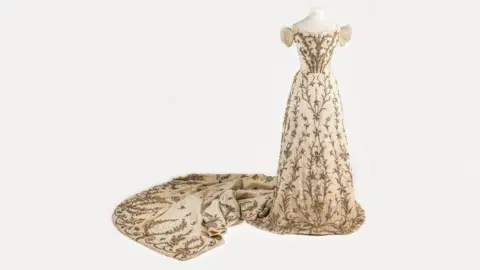 Fashion Museum Bath A cream silk court dress by Worth on display in Paris. It has a train with diamantes and all over silver and gold metal thread zardozi embroidery. It is seen on a white mannequin with a white background.