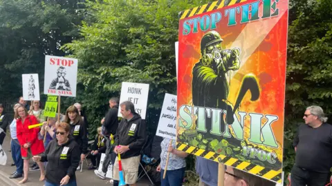 Protestors lined up on a pavement next to some trees, some holding signs that say "stop the stink".