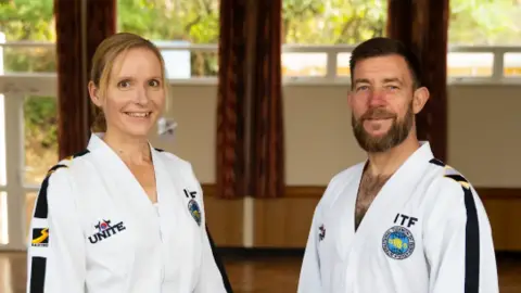 A man and a woman are pictured in white taekwondo clothing with blue stripes down their arms. The woman, positioned on the left, has tied back blonde hair and brown eyes. The man, on the right, has short brown hair and a brown beard with blue eyes.