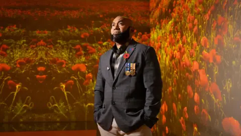 Royal British Legion A man in a navy suit, beige trousers and is standing in a room with red poppies wall paper. He has military medals attached to his suit.