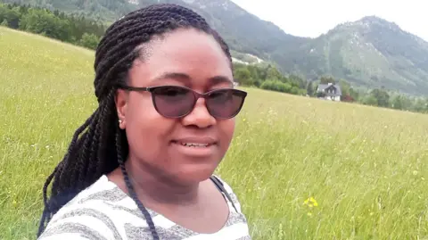 A young black woman wearing sunglasses with her hair in braids, posing for a selfie in a meadow with hills in the background